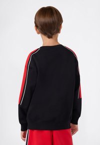 Bambino con capelli corti castani, che indossa una felpa nera con strisce rosse e bianche sulle maniche e pantaloni corti rossi, di spalle rispetto all'osservatore.