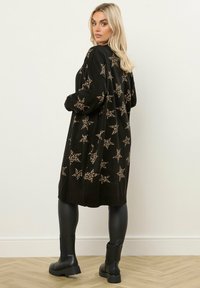 Czarny kardigan z dużymi gwiazdami w leoparda. Długie rękawy, luźny krój, miękka faktura. Połączony z czarnymi legginsami i masywnymi czarnymi butami.
