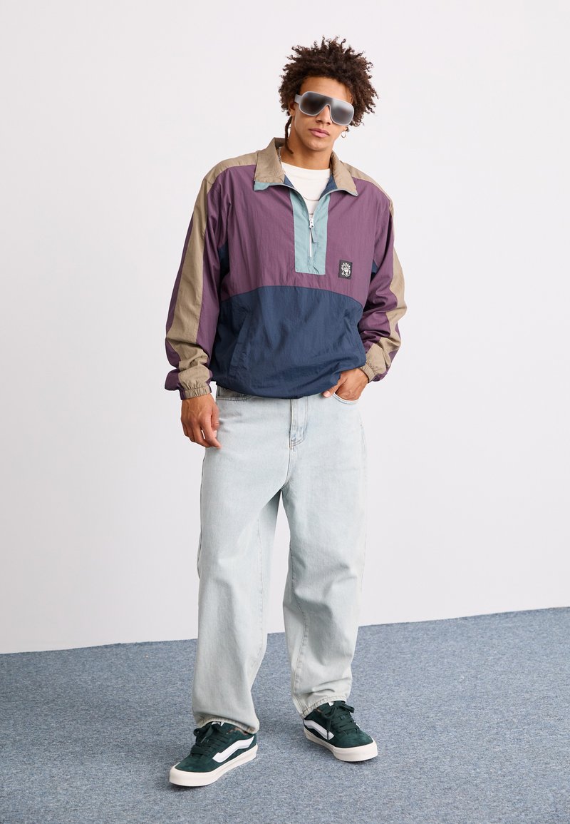 Veste coupe-vent en violet, marine et beige avec une fermeture éclair quart et des poignets élastiques. Associée à un jean clair et des baskets en daim vert.