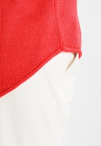 Czerwony sweter wełniany o teksturowym wykończeniu, z ukośnym dołem. Noszony na kremowych spodniach, podkreślających detal kieszeni.