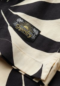Tela con un patrón en blanco y negro que muestra un diseño de hojas. Una etiqueta negra con bordado dorado presenta "Superdry" sobre un sol y palmeras.