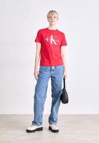 Rode katoenen T-shirt met witte "Calvin Klein Jeans" logo, blauwe jeans, zwarte handtas en crèmekleurige schoenen met stevige zool op een neutrale achtergrond.