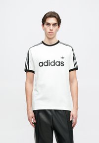 Biela bavlnená tričko s čiernym rebrovaným golierom, krátkymi rukávmi a tromi čiernymi pruhmi na pleciach. Čierne logo "adidas" je umiestnené na prednej strane.