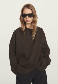 Frau mit langen blonden Haaren, die einen schwarzen oversized Pullover, schwarze Hosen und schwarze SonnenbrILLE trägt und vor einem schlichten hellen Hintergrund posiert.