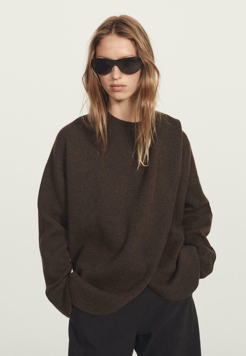 Woman with long blonde hair wearing black oversized sweater, black pants, and black sunglasses, posing against plain light background.