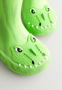 Bottes de pluie en caoutchouc vertes avec un design texturé en crocodile, de grands yeux et des crocs blancs à l'avant. Surface lisse et semelles épaisses.