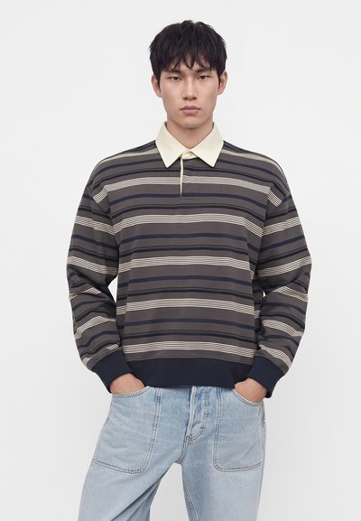 Jeune homme portant un maillot de rugby rayé à manches longues avec un col beige et un jean bleu clair, les mains dans les poches, expression neutre.