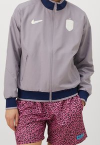 Gray zip-up jacket with navy collar, features Nike logo; paired with pink shorts in a black leopard print, accentuated by a blue detail.