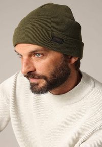 Bonnet en tricot côtelé vert olive avec une étiquette noire, porté par une personne. La personne porte un pull texturé de couleur claire.