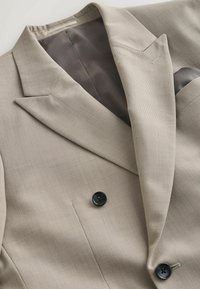 Blazer beige sur mesure avec revers crantés, boutons sombres et doublure intérieure foncée, posé à plat sur une surface claire.