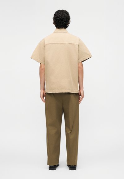 Άνδρας με κοντά σγουρά μαλλιά στέκεται στραμμένος προς τα πίσω, φοράει μπεζ πουκάμισο με κοντά μανίκια και παντελόνι ελιάς με μαύρα παπούτσια.