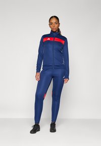 Tuta atletica blu navy con giacca con zip, caratterizzata da dettagli rossi e logo, pantaloni aderenti con dettagli rossi, texture liscia e materiale elasticizzato.