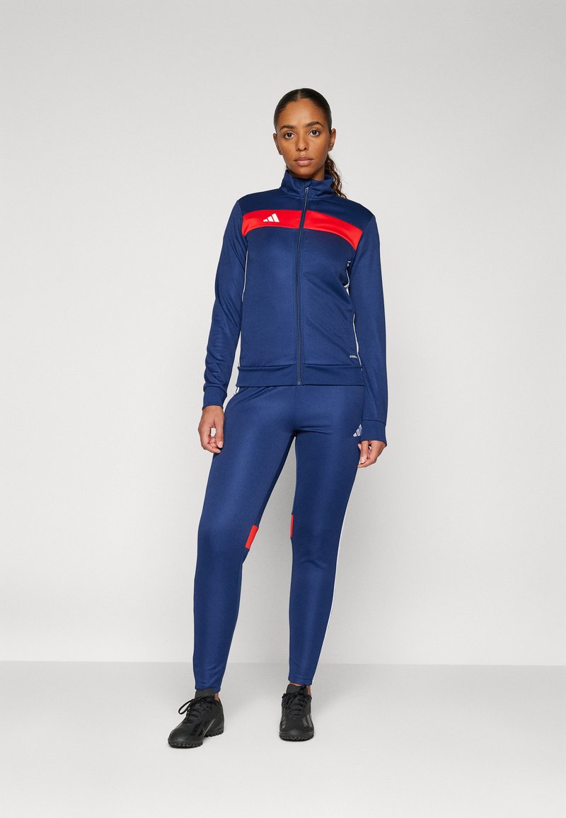 Tuta atletica blu navy con giacca con zip, caratterizzata da dettagli rossi e logo, pantaloni aderenti con dettagli rossi, texture liscia e materiale elasticizzato.