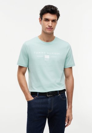 Mann med mørkt krøllete hår iført en lyseblå kortermet Tommy Hilfiger t-skjorte, mørke jeans og svart belte, stående med én hånd i lommen.