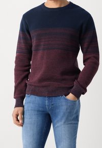 Hombre vistiendo un suéter de punto navy y burdeos con rayas horizontales y jeans de mezclilla azul claro, de pie con una mano en el bolsillo.