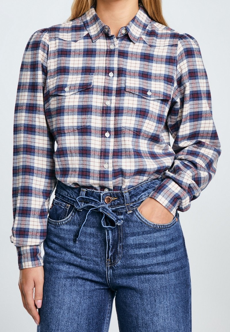Femme portant une chemise à carreaux bleue et blanche boutonnée, rentrée dans un jean bleu taille haute avec une ceinture en tissu nouée.