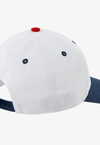 Casquette de baseball avec un corps en coton blanc, une visière bleu marine et un bouton rouge sur le dessus. Comprend des œillets de ventilation sur les côtés.