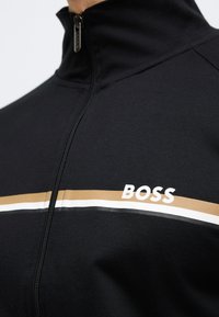 Μαύρο μπουφάν με φερμουάρ, διαθέτει όρθιο γιακά, οριζόντιες λευκές και χρυσές ρίγες, με το λογότυπο "BOSS" σε λευκό στην αριστερή πλευρά.