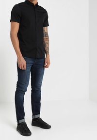 Svart kortärmad skjorta med krage, matchad med mörkblå jeans och svarta sneakers, som visar en slimmad passform och avslappnad design.