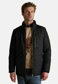 Czarna pikowana kurtka z kołnierzem stojącym, z zamykanymi na zamek kieszeniami, noszona na beżowym swetrze z haftowanym wzorem lwa.