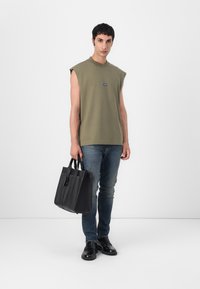 Camisa sin mangas de color verde oscuro, parche de logo azul, combinada con jeans azules y zapatos de cuero negros, sosteniendo una bolsa de tote negra con asas dobles.