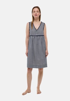 Femme debout pieds nus portant une robe sans manches à carreaux noirs et blancs, longueur genoux, avec des boutons, sur un fond uni.