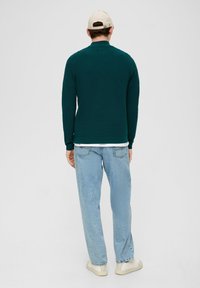Türkiser gestrickter Pullover mit Kragen, kombiniert mit hellblauen, locker sitzenden Jeans. Das Outfit zeichnet sich durch eine dezente Textur und eine klassische Silhouette aus.
