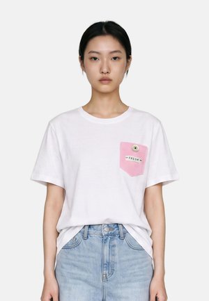 Giovane donna che indossa una t-shirt bianca con una tasca rosa etichettata "FRESH" e jeans a vita alta azzurri chiaro su uno sfondo semplice.