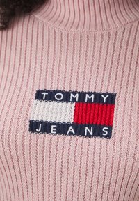 Rosa ribbad polokrage-tröja med en rektangulär broderad logotyp i marinblått, vitt och rött med texten "TOMMY JEANS" nedanför.