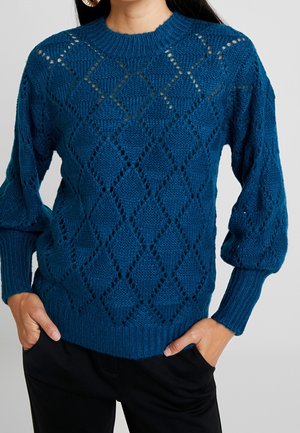 Femme portant un pull en maille bleu avec un motif losange, poignets et ourlet côtelés, mains dans les poches, pantalon noir visible en dessous.