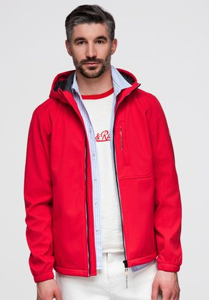 Homme aux cheveux gris courts et à la barbe portant une veste rouge ouverte sur une chemise blanche et une chemise bleu clair à boutons sur un fond uni.