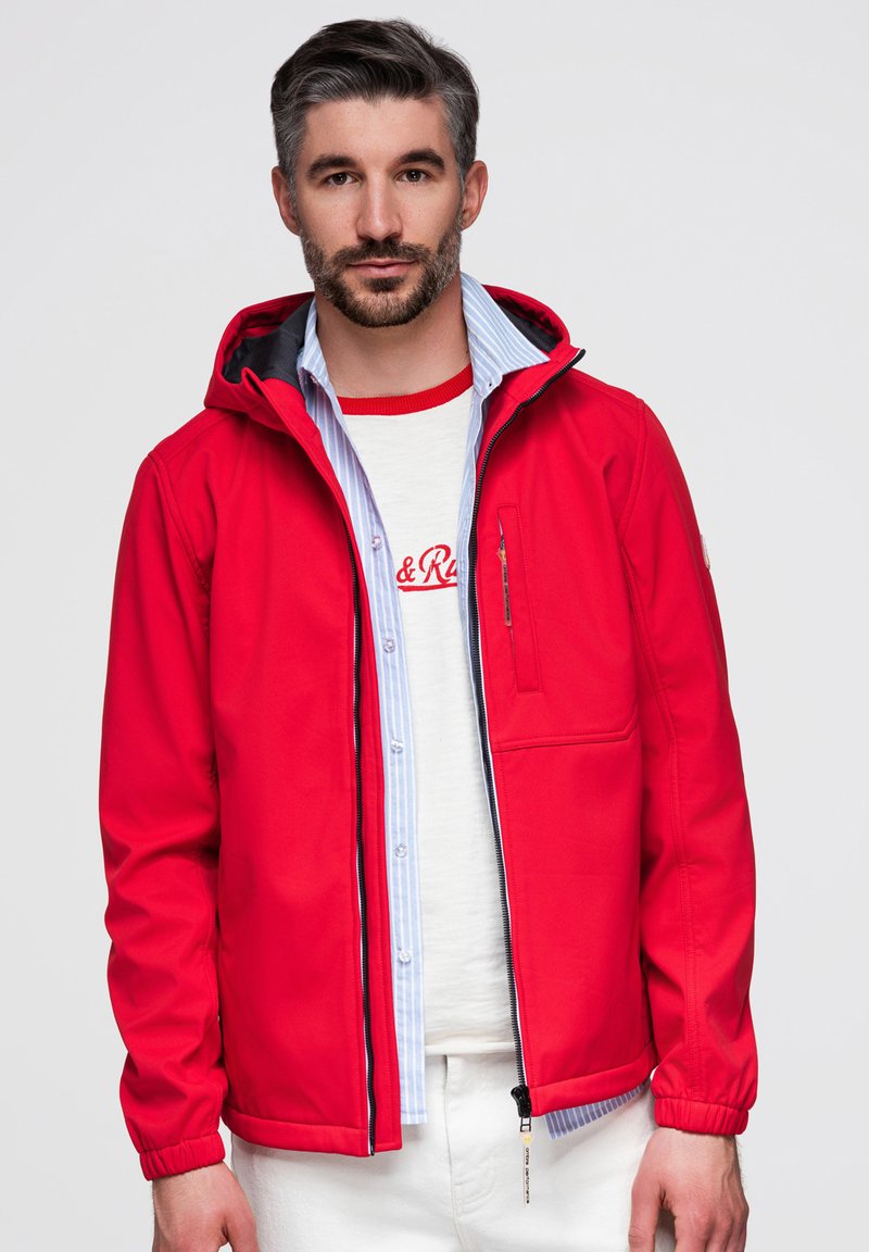Homme aux cheveux gris courts et à la barbe portant une veste rouge ouverte sur une chemise blanche et une chemise bleu clair à boutons sur un fond uni.