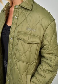 Chaqueta acolchada color verde oliva con cierre de botones a presión, gran bolsillo frontal y logotipo bordado en el pecho. Textura suave y acolchada.