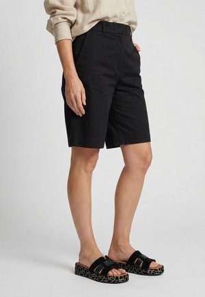 Person iført beige langærmet skjorte, sorte knælange shorts og sorte platformsandaler med mønstrede såler, stående mod en ensfarvet baggrund.