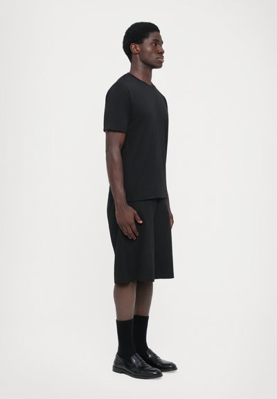 Schwarzes T-Shirt mit kurzen Ärmeln und knielangen Shorts, beide aus glattem Stoff. Kombiniert mit schwarzen Socken und Slippern, vor einem weißen Hintergrund stehend.