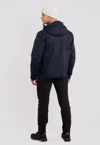Marineblauwe waterdichte jas met een capuchon, verstelbare manchetten, ritszakken en een slank ontwerp, gecombineerd met zwarte broek en donkere schoenen.
