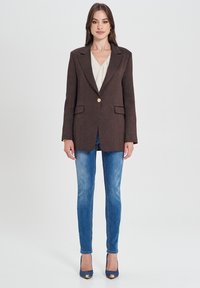 Blazer marrone con texture, dotato di un singolo bottone dorato, abbinato a jeans skinny blu chiaro e tacchi blu con accenti metallici.