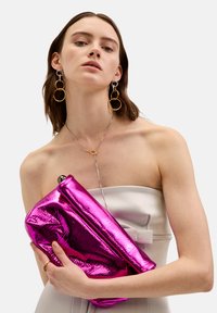 Fuchsiová metalická kabelka s měkkou, lesklou texturou a skládacím designem, držena proti svetle zbarvenému, bezramennému topu.
