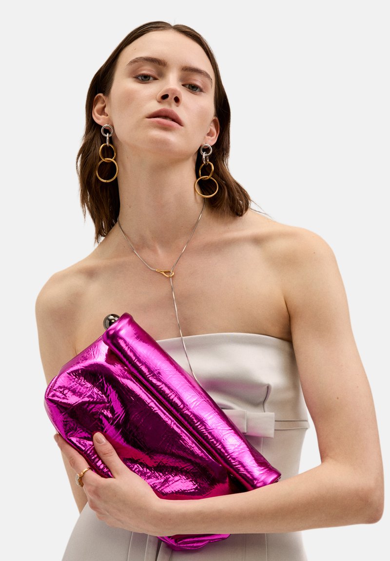 Fuchsiová metalická kabelka s měkkou, lesklou texturou a skládacím designem, držena proti svetle zbarvenému, bezramennému topu.