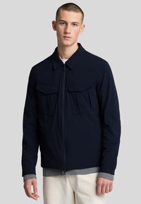 Giacca navy con zip frontale, colletto piatto, due tasche sul petto e un maglione grigio a coste sotto. Tessuto morbido con taglio moderno.