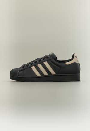 Baskets Adidas Superstar noires avec des rayures beige, des lacets noirs et un arrière beige sur un fond neutre.