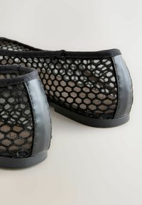 Černé síťované tenisky s hexagonálním otevřeným vzorem, pevným černým lemováním a patou, zobrazené na jednoduchém světlém pozadí.