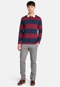 Polo a manica lunga con righe orizzontali blu navy e bordeaux, colletto bianco, abbinato a pantaloni grigi e scarpe da barca marroni.