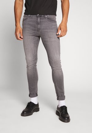 Vaqueros de mezclilla gris con corte ajustado, que cuentan con un diseño clásico de cinco bolsillos y bajos remangados, combinados con zapatos negros y calcetines blancos.