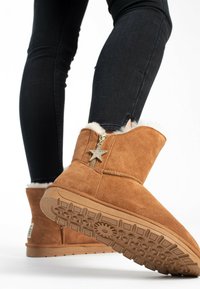 Botas de tobillo de ante en color beige con un forro blanco esponjoso, una cremallera lateral con un charm de estrella y una suela de goma texturizada.