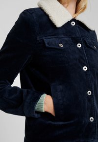 Marinblå kordjacka med en vit fuskpälskrage, silverfärgad hårdvara, två bröstfickor och en synlig grön stickad mudd vid ärmen.