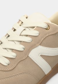 Μπεζ αθλητικό παπούτσι με υφασμάτινο επάνω μέρος, λευκές δερμάτινες λεπτομέρειες, επίπεδα κρεμ κορδόνια και σόλα από καουτσούκ με καφέ απόχρωση, διαθέτοντας καμπυλωτή ραφή σχέδιο.