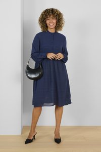 Abito azzurro navy con bottoni frontali, righe texturizzate, maniche lunghe, vita arricciata e una vestibilità comoda. Abbinato a una borsa nera e tacchi.
