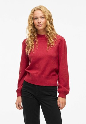 Rød strikket sweater med høj hals, lange ærmer med let puff, og en tætsiddende talje. Bæres med sorte bukser.
