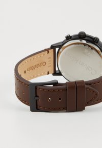 Bracelet de montre en cuir marron avec des coutures contrastées, hardware en métal noir et dos en argent poli. Comprend des perforations pour un ajustement.